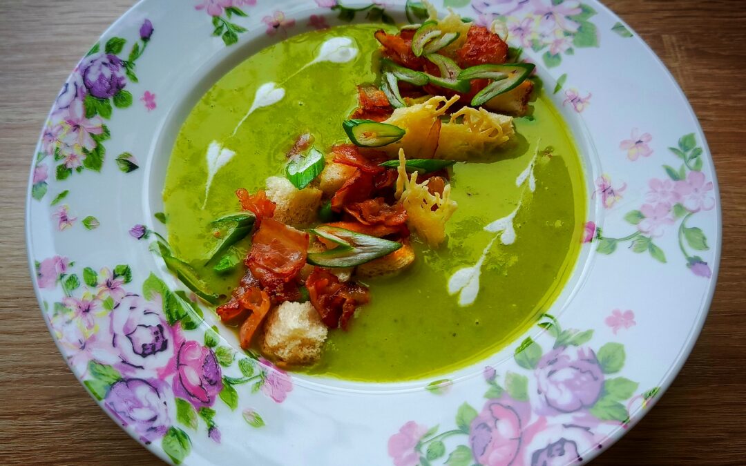 hrášková polévka s krutony, slaninou, sýrovým chipsem, zdobená smetanou ve tvaru srdíček. Vše na talíři s květinami